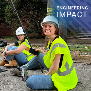 Two people wearing safety vests and hard hats sit on the ground