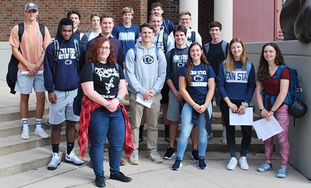 A group of students with their teacher stand together and smile on a sunny day in front of a brick building.