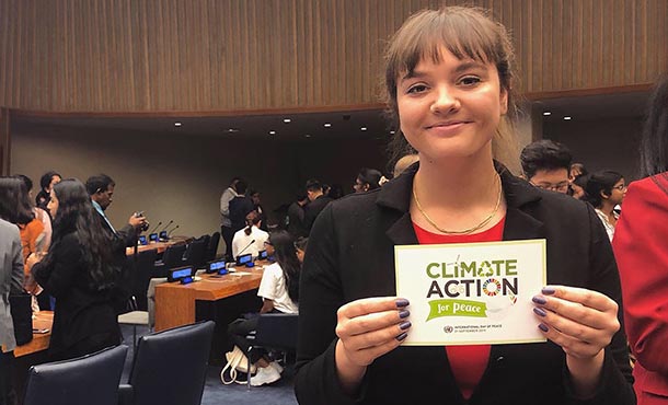 A smiling woman stands in a crowded room, with tables, chairs and other people visible in the background. She holds up a Climate Action for Peace sign.