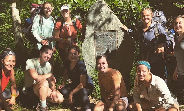 Nine women dressed in hiking gear pose for a photo near a rock