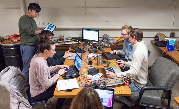 Six students sit with their laptops around a table, which is covered with wires and electronic components.