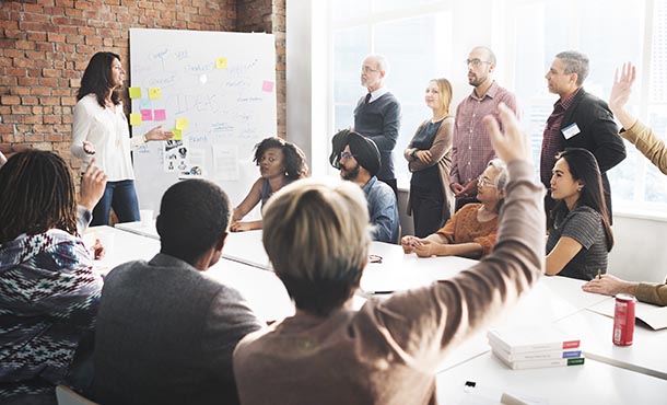 A dozen people sit and stand around a table as a woman leads a discussion at the front of the room