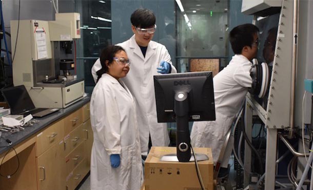 Two men and a woman in lab coats looking at polymer samples via a glove box and monitor.