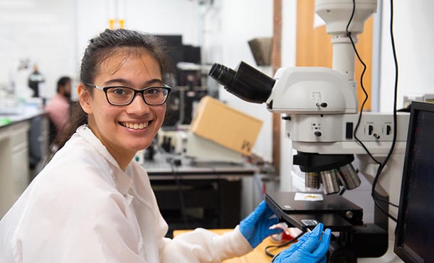 A researcher smiles at the camera while sitting in front of a microscope/A researcher works at a computer