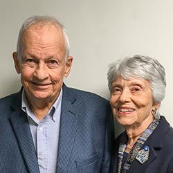 A man wearing a button-down shirt and suit jacket and a woman wearing a dark top with a brooch smile at the camera in front of a beige wall.