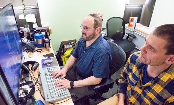 A man in a blue shirt with glasses and a man in a plaid shirt to his right work at a computer