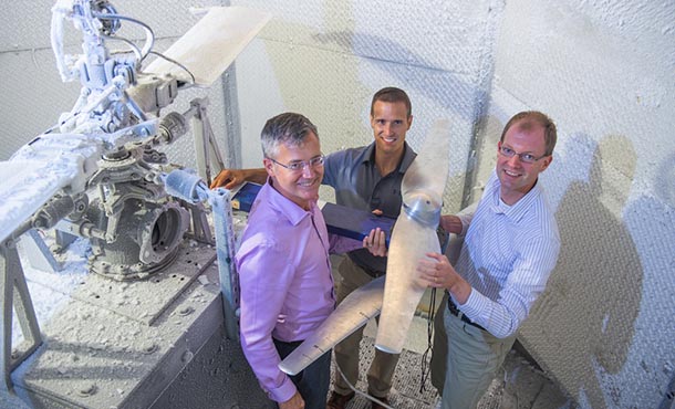 Three men stand in a steel chamber, around a steel blade and next to a larger rotor. The rotor and the walls of the chamber are covered in ice.