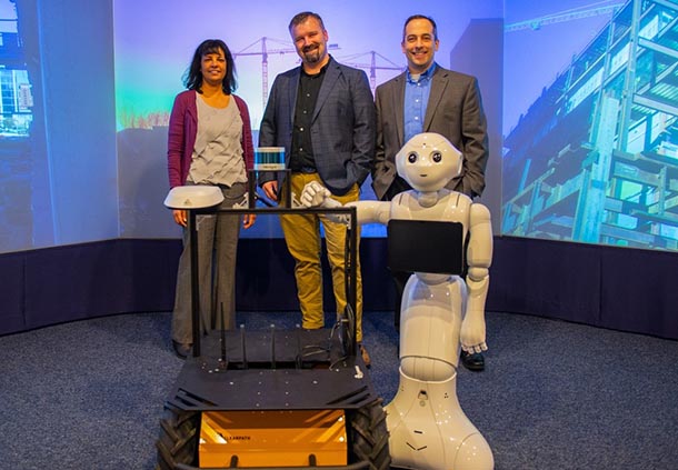 one woman and two men stand behind two medium-sized robots