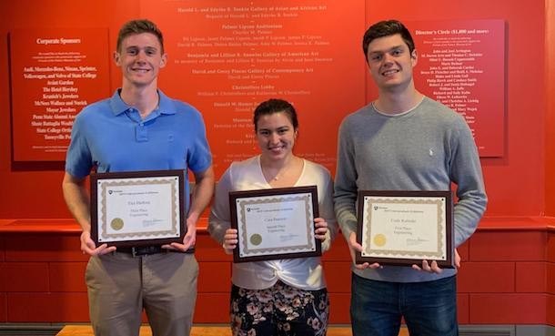 Three students, two male, one female, each holding framed Undergraduate Exhibition award in front of orange background.