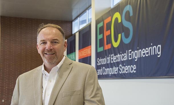 A man in a tan suit jacket poses in front of a School of E E C S wall banner