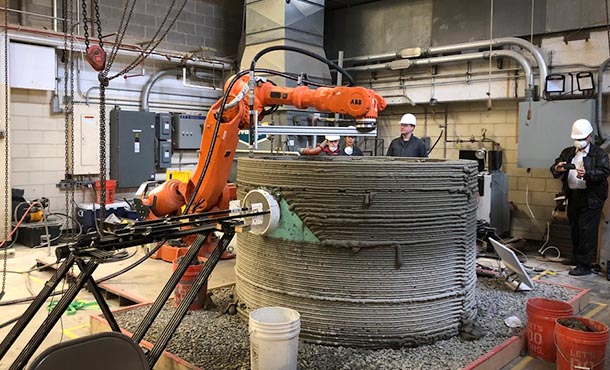 Four people look at an orange robotic arm as it moves above a cylinder concrete structure