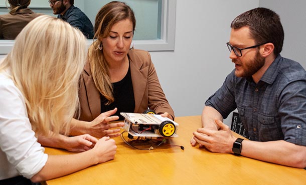 Two women and one man sit around a table looking at a robotic car built from poster foamboard, wheels, batteries and wires.