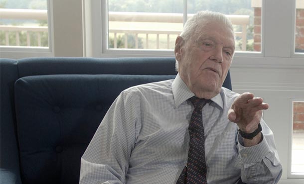 elderly man wearing a shirt and tie sitting in a chair speaking