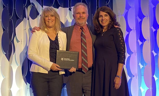 Two women stand on either side of a man as they hold a Penn State Alumni Association award.