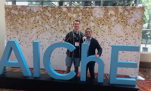 Two men standing in front of large letters spelling out A I C H E