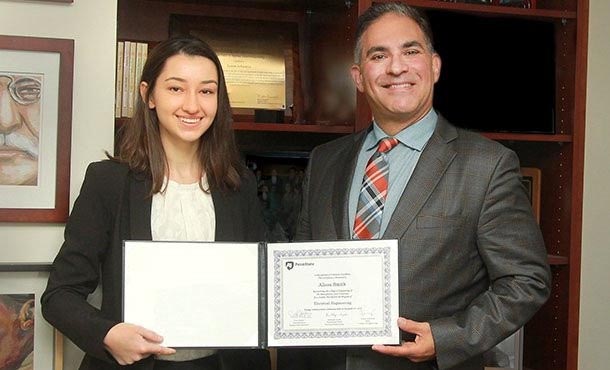 A woman in a suit jacket and a man in a suit smile and pose with a certificate