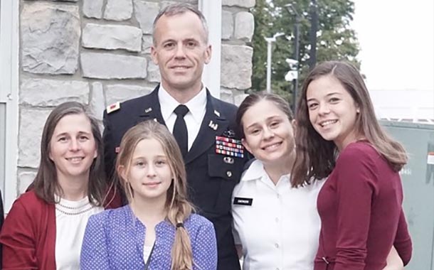 Man dressed in a military uniform stands on a porch with his wife and three daughters