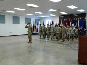 Saluting military man, dressed in uniform, stands in large room with group of soldiers in line behind him