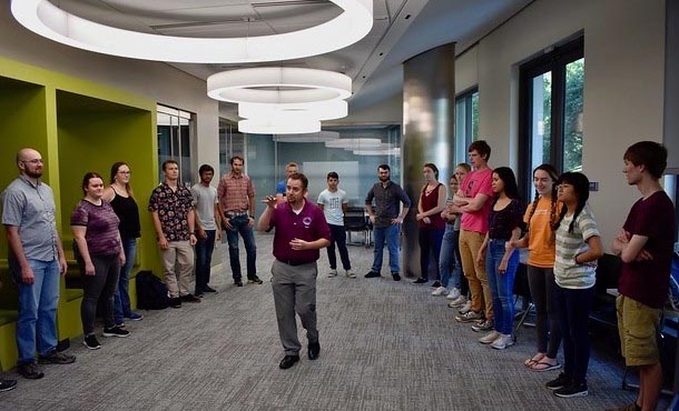 Man in red shirt standing in center of circle of students and gesturing with hands.