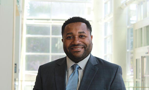 A headshot of Kwesi Vincent smiling at the camera and wearing a gray suit jacket, white shirt and light blue tie