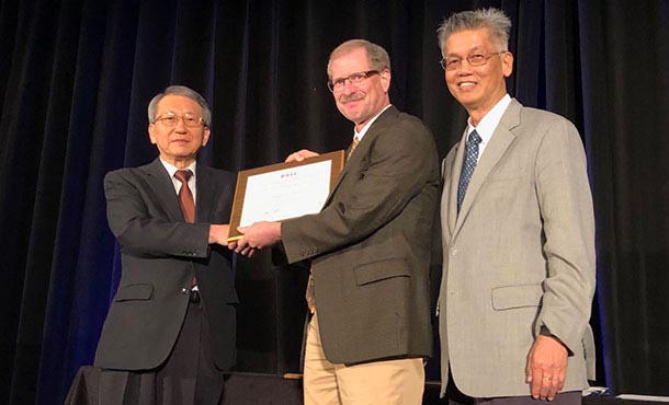 Two men shake hands as one man hands the other man a plaque. A third man stands by.