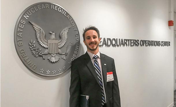 A man in a suit smiles and stands in front of a seal that reads Unites States Nuclear Regulatory Commission.