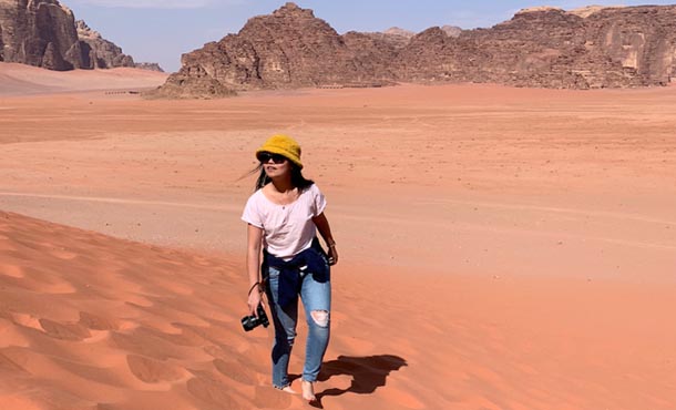 Young woman wearing sunglasses, a yellow hat, white t-shirt and jeans walks through the desert with a camera and stares off into the distance.