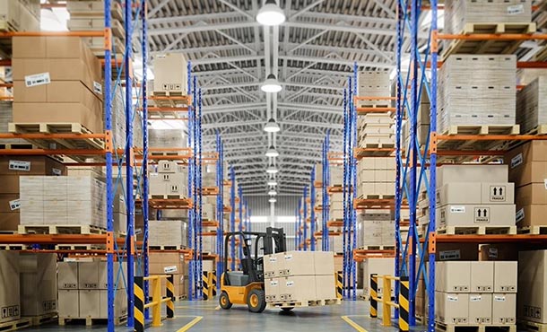 Forklist in warehouse aisle loaded with a box. Along the aisle are stacks of boxes for distribution.