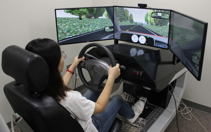 A student demonstrates a driving simulator that consists of a car seat, a wheel, and three screens showing a road on them. 