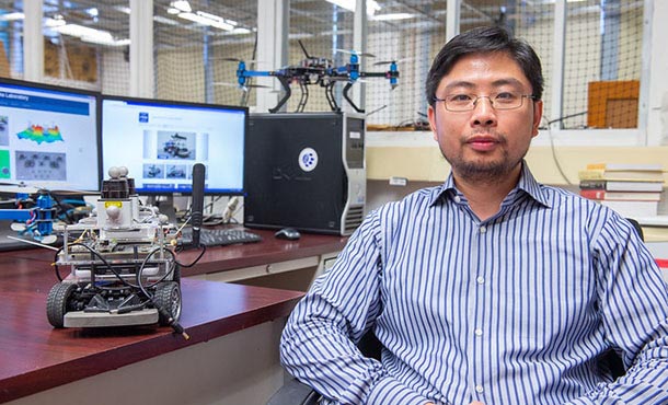 Minghui Zhu sits at his desk. On the desk is a small device with wheels and wires, and behind him on the desk are two computer monitors and another small robot-like device.