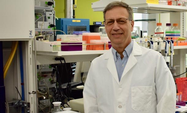 Man with glasses wearing white lab coat and standing in lab with lab equipment in background.